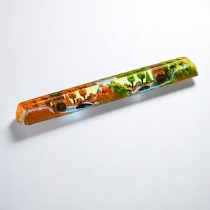 Może przedstawiać: Przezroczysty klawisz z żywicy z kolorowym wzorem krajobrazu przedstawiającym drzewa, wodę i skały. Klawisz jest prostokątny i ma gładkie, błyszczące wykończenie.