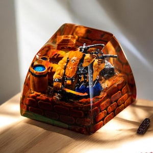 Op de afbeelding: Een transparante hars toetsenkap met een gedetailleerd miniatuur-tafereel van een krijgerfiguur dat een wapen vasthoudt, staand op een bakstenen muur met een vat en andere details. De scène is ingebed in de toetsenkap, waardoor een 3D-effect ontstaat.