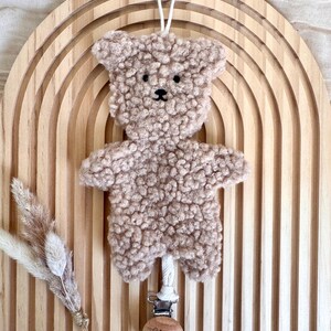 Könnte beinhalten: Ein kuscheliger, hellbrauner Teddybär mit schwarzer Nase und Augen, an einer weißen Schnur aufgehängt. Der Bär ist an einem hölzernen Schnullerclip mit dem eingravierten Namen "Elias" befestigt. Der Bär befindet sich vor einem hölzernen Regenbogenhintergrund.