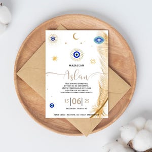 May include: A white invitation card with gold accents, featuring an eye symbol, crescent moon, and sunburst designs. The card includes text in a script font and is placed on a wooden plate with a brown envelope.