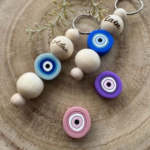 Könnte beinhalten: Zwei Schlüsselanhänger aus Holzperlen mit den eingravierten Namen "Aslan" und "Levin". Jeder Schlüsselanhänger hat einen blauen bösen Blick-Anhänger und eine Holzperle. Es gibt auch zwei weitere böse Blick-Anhänger, einen in Lila und einen in Rosa.