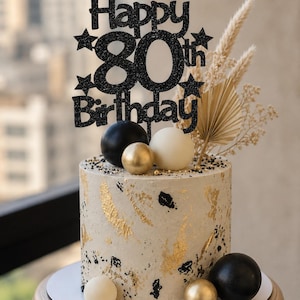 Op de afbeelding: Een verjaardagstaart versierd met een zwarte glitter "Happy 80th Birthday" taarttopper. De taart is wit met gouden en zwarte accenten, en gegarneerd met zwarte, witte en gouden bollen en gedroogde bloemelementen.