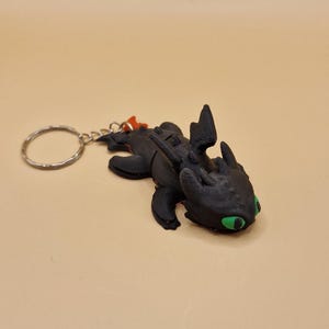 May include: Black dragon keychain with green eyes and an orange tail fin. The dragon is attached to a silver key ring.