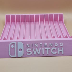 May include: A pink Nintendo Switch game cartridge holder. The holder has a series of vertical slots and the Nintendo Switch logo in white. The holder is made of plastic.