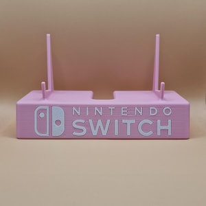 May include: A light pink Nintendo Switch stand with the Nintendo Switch logo and text in white. The stand has two vertical supports and a rectangular base. The stand is designed to hold a Nintendo Switch console.