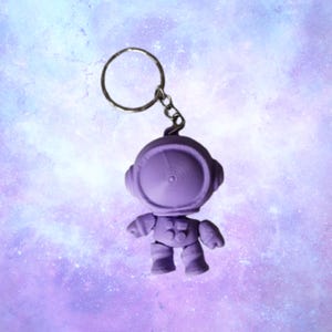 May include: A purple astronaut keychain with a silver keyring and chain. The astronaut figure has a detailed helmet, arms, and legs. The background is a soft purple and blue galaxy design.