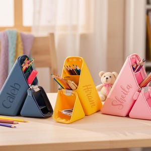 Puede incluir: Tres estuches personalizados en azul, amarillo y rosa, cada uno con un nombre impreso en el lateral. Los estuches son triangulares y se abren para revelar compartimentos llenos de lápices y artículos de papelería. Un osito de peluche está detrás de los estuches amarillos y rosas.