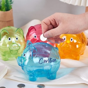 Op de afbeelding: Vier doorzichtige plastic spaarpotten in verschillende kleuren: groen, rood, oranje en blauw. De blauwe spaarpot heeft de naam "Carlton" erop.