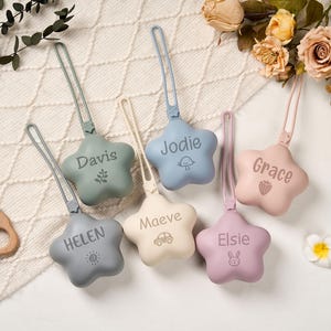 May include: Six star-shaped baby pacifier holders in pastel shades of sage green, light blue, cream, grey, and pink. Each has a name and a small icon engraved. A wooden teething ring is also visible.