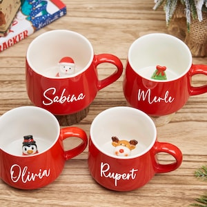 Puede incluir: Cuatro tazas rojas de cerámica con interiores blancos, cada una con una figura diferente con temática navideña en su interior. Las tazas tienen nombres escritos: Sabina, Merle, Olivia y Rupert. Las tazas están sobre una superficie de madera.