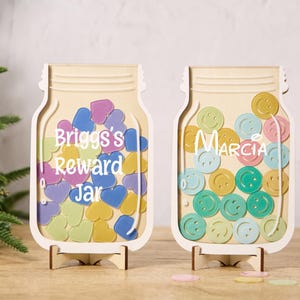 以下が含まれることがあります： スタンド付きの木製のメイソンジャー型の報酬チャート2つ。左側のチャートは、薄い青、黄色、ピンクのハートのデザインで、「Briggs's Reward Jar」と書いてあります。右側のチャートは、薄い青、黄色、緑のスマイリーフェイスのデザインで、「Marcia」と書いてあります。