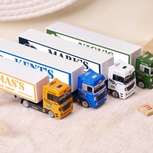 Peut inclure: Plusieurs petits camions jouets de couleurs vives, dont jaune, bleu, blanc et vert. Chaque camion est accompagné d'une remorque blanche portant des noms d'entreprises. Les jouets sont disposés sur une surface claire.
