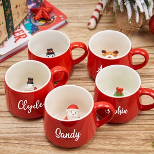 Könnte beinhalten: Fünf rote Keramikbecher mit weißen Innenräumen, jeder mit einer anderen weihnachtlichen Figur im Inneren. Auf den Bechern stehen Namen. Ein Schneemann, ein Pinguin, ein Rentier, der Weihnachtsmann und ein Weihnachtsbaum sind die Figuren.