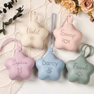 Può includere: Cinque porta-ciuccio a forma di stella in colori pastello: beige, rosa, azzurro, lavanda e verde salvia. Ognuno ha un nome inciso: Violet, Sandy, Darcy, Sophia e Frank. Hanno un anello per attaccare il ciuccio.