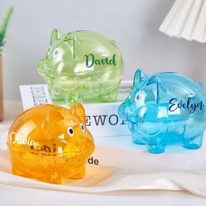 Puede incluir: Tres alcancías de plástico transparente con nombres personalizados. Las alcancías son de color verde, azul y naranja. Los nombres en las alcancías son David, Evelyn y Olivia.