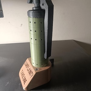 Könnte beinhalten: Ein 3D-gedrucktes Objekt in Form einer Granate in Grün und Schwarz mit einem Holzsockel. Der Sockel trägt die Gravur "Complaint? Take One".