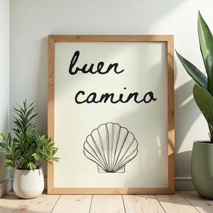 May include: A framed print with the words "buen camino" and a seashell illustration. The artwork is in a light wood frame, with a cream-colored background. The print is displayed on a wooden floor, with potted plants on either side.