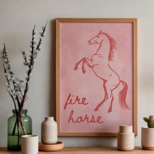 May include: A framed print with a rearing horse silhouette in reddish-orange on a pink background. The words "fire horse" are written below the horse. The frame is made of light-coloured wood.