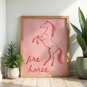 May include: A framed print with a coral-coloured illustration of a rearing horse on a pink background. The words "fire horse" are written in a cursive font below the horse. The frame is made of light-coloured wood.
