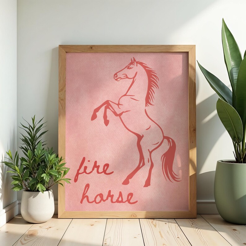 May include: A framed print with a coral-coloured illustration of a rearing horse on a pink background. The words "fire horse" are written in a cursive font below the horse. The frame is made of light-coloured wood.