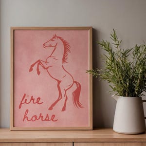 May include: A framed print with a reddish-orange horse illustration on a pink background. The words "fire horse" are written below the horse. The frame is made of light-coloured wood. The print is displayed on a wooden surface.