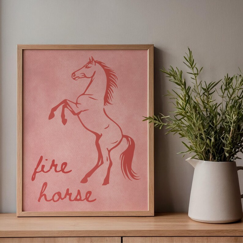 May include: A framed print with a reddish-orange horse illustration on a pink background. The words "fire horse" are written below the horse. The frame is made of light-coloured wood. The print is displayed on a wooden surface.