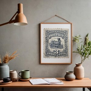 May include: Framed vintage postage stamp print featuring a detailed illustration of a steam train. The stamp is in shades of blue and white, with the text "UNITED STATES POSTAGE" and "THREE CENTS". The print is displayed in a wooden frame, hanging on a gray wall.