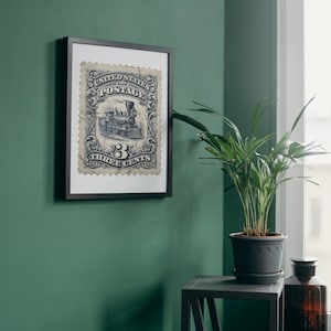 May include: Framed vintage postage stamp art featuring a detailed illustration of a steam train. The stamp is in shades of blue and white, with the text "UNITED STATES POSTAGE" and "THREE CENTS". The artwork is displayed in a black frame against a green wall.