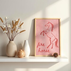 May include: A framed print with a red horse illustration on a pink backdrop, with the words "fire horse" beneath. The print is displayed on a white shelf, alongside vases and decorative items, creating a stylish home decor piece.
