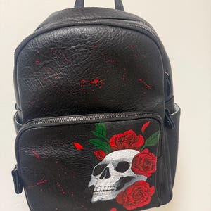 Può includere: Zaino nero con schizzi di vernice rossa e un teschio dipinto con rose rosse. Lo zaino ha una tasca frontale e due tasche laterali. La borsa è tenuta da una persona.