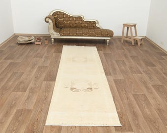 May include: A long, rectangular, cream-colored rug with a central floral design. The rug is placed on a hardwood floor, with a decorative chaise lounge and small wooden stool in the background. The rug's dimensions are approximately 3 x 8 feet.