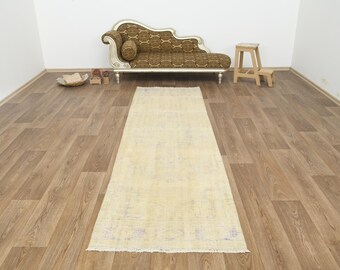 Puede incluir: Una alfombra rectangular de color claro con un aspecto descolorido y vintage. Está colocada sobre un suelo de madera, con un chaise longue estampado y un pequeño taburete de madera en el fondo. La alfombra mide unos 200 cm de largo.