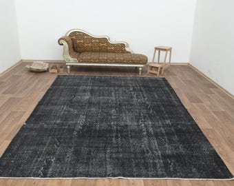 Op de afbeelding: Een groot, rechthoekig, verweerd zwart tapijt met witte accenten. Het tapijt ligt op een houten vloer. Een decoratieve chaise longue en een kleine houten kruk staan op de achtergrond.