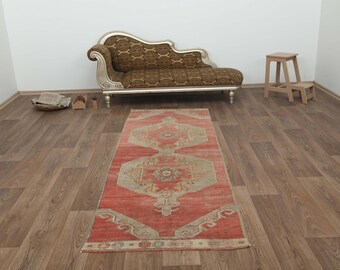 May include: A red and beige patterned runner rug with a geometric design. The rug is laid out on a wooden floor in a room with a chaise lounge and a small wooden stool.
