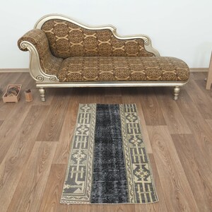 May include: A rectangular, vintage-style rug with a central dark blue section and patterned borders in beige and gray. The rug is placed on a wooden floor, with a chaise lounge and small wooden stool in the background.