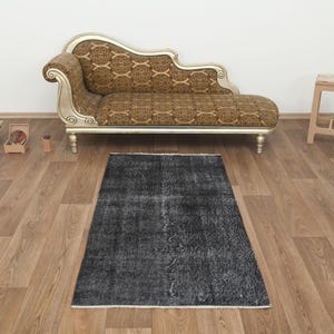 Può includere: Un tappeto rettangolare grigio scuro dall'aspetto vissuto. Il tappeto è posizionato su un pavimento in legno, con una chaise longue decorativa e uno sgabello in legno sullo sfondo. Le dimensioni del tappeto sono di circa 90 x 150 cm.