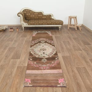 May include: A long, rectangular runner rug featuring a deep brown base color with a central cream-colored medallion. The rug is decorated with intricate floral patterns in shades of pink and white, with a faded, distressed texture throughout the fabric.