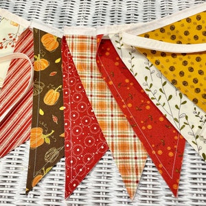May include: A fall-themed fabric banner with nine triangular flags. The flags are made of different fabrics with various autumnal patterns, including pumpkins, leaves, and berries. The flags are connected by a white string.