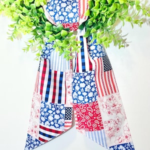 May include: A patriotic wreath accent featuring a patchwork design in red, white, and blue. The fabric includes floral, paisley, striped, and American flag patterns. The wreath is adorned with vibrant green foliage.