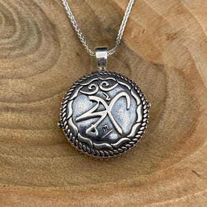 Op de afbeelding: Een zilverkleurige ketting met hanger. De ronde hanger heeft een ingewikkeld ontwerp binnen een touwachtige rand. De ketting is ook zilverkleurig en hangt tegen een houten achtergrond.