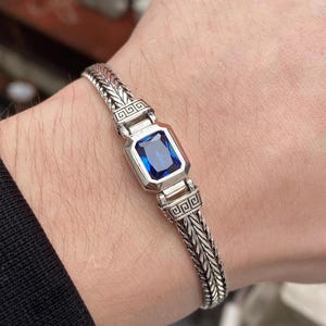 May include: Silver bracelet with a rectangular, deep blue gemstone. The bracelet has a woven chain design with geometric accents. The jewellery is displayed on a wrist, highlighting the bracelet's details and craftsmanship.