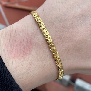 Puede incluir: Una pulsera dorada con un diseño de cadena detallado. La pulsera se lleva en la muñeca, mostrando sus intrincados eslabones y su superficie brillante y reflectante. La joya es un accesorio clásico.