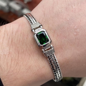 Puede incluir: Una pulsera de plata con un diseño de cadena trenzada. La pulsera presenta una piedra preciosa verde esmeralda rectangular engastada en un bisel de plata. La pulsera tiene patrones geométricos decorativos.