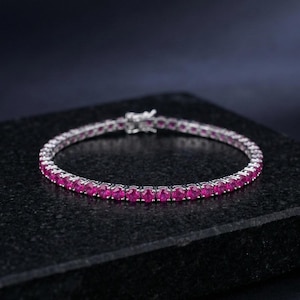 Puede incluir: Una pulsera plateada con una fila de gemas rosas redondas y vibrantes. La pulsera se muestra sobre un fondo oscuro, resaltando el brillo de las gemas. El cierre es visible, lo que indica el diseño y la funcionalidad de la pulsera.