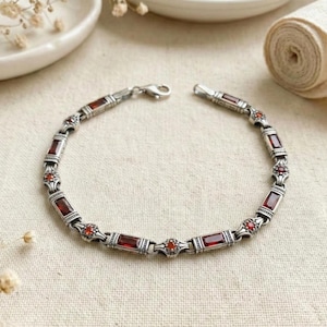 May include: Silver bracelet featuring rectangular and round red gemstones. The bracelet has an ornate design with a lobster clasp closure. The gemstones are set in a silver metal setting. The bracelet is displayed on a neutral-colored surface.