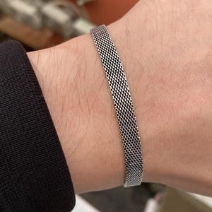 Può includere: Un braccialetto a rete argentata è indossato su un polso. Il braccialetto è realizzato in metallo fine e intrecciato, creando una superficie strutturata. Il braccialetto è un design semplice ed elegante, adatto all'uso quotidiano. Lo sfondo è sfocato, con una manica nera visibile.
