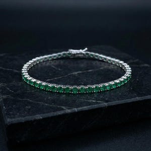 May include: A silver bracelet featuring a row of vibrant green emeralds. The gemstones are set closely together, creating a continuous line of color. The bracelet is displayed on a dark, textured surface.