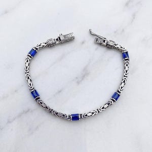 May include: Silver-toned bracelet with a Byzantine chain design, featuring rectangular blue gemstone accents. The bracelet is laid out on a white marble surface, showcasing its intricate details and clasp mechanism. A stylish piece of jewelry.