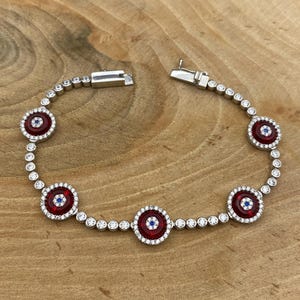 May include: A silver bracelet with a series of red and white circular charms. Each charm has a red center with a blue accent and is surrounded by small white stones. The bracelet is on a wooden surface.