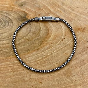 Könnte beinhalten: Ein silberfarbenes Armband mit Perlenmuster. Das Armband hat einen sicheren Verschluss und wird auf einer Holzoberfläche gezeigt. Die Perlenkette besteht aus kleinen, runden Perlen. Dieses Schmuckstück ist ein stilvolles Accessoire.